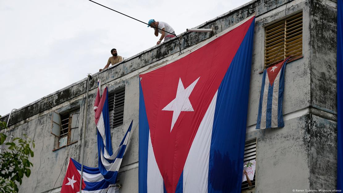 Homens penduram bandeiras cubanas sobre as janelas da casa do ativista da oposição Yunior García Aguilera, numa tentativa de impedi-lo de se comunicar com o mundo exterior, enquanto ele segura uma flor com o braço estendido para fora da janela.