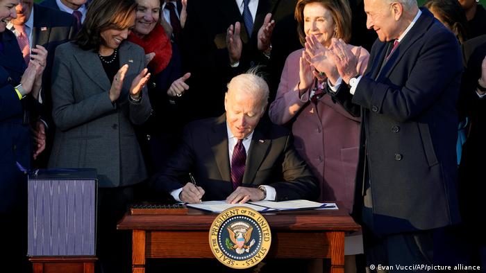 Biden sitzt an seinem Pult und unterzeichnet das Gesetz, um ihn stehen klatschend seine Parteifreunde und Mitarbeitenden. 