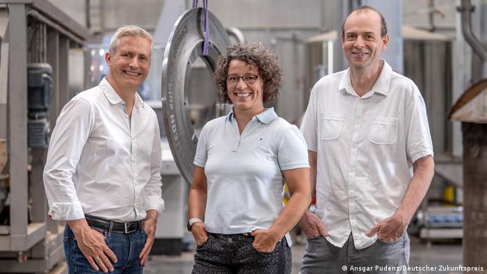 Deutscher Zukunftspreis 2021: In der Reifenproduktion von Continental bei Hannover stehen Dirk Prüfer, Carla Recker und Christian Schulze-Gronover. Im Hintergrund eine Reifen-Schablone aus Metall. 
