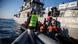 A coast guard boat and two small rubber boats, filled with people in life vests and an aid worker with NGO Emergency Response Centre International in a yellow vest A coast guard boat and two small rubber boats, filled with people in life vests and an aid worker with NGO Emergency Response Centre International in a yellow vest