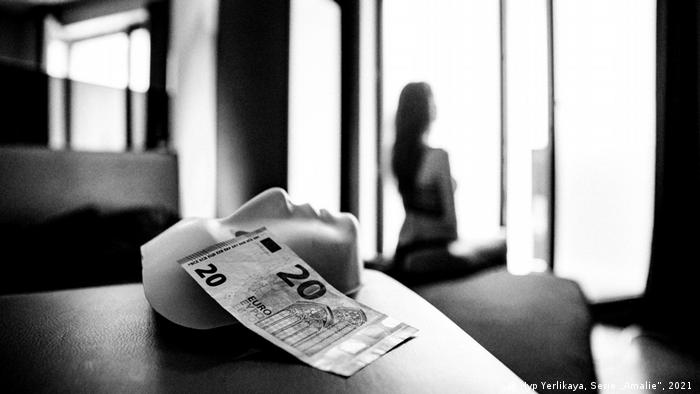 Eine Frau sitzt leicht bekleidet auf einem Bett. Auf einem Tisch liegen eine Gesichtsmaske und ein Geldschein. 