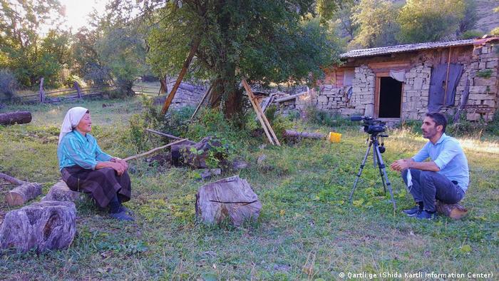 Journalist Saba Tsitsikashvili interviewing a villager on the border with South Ossetia: they are sitting outside on tree stumps in front of a stone hut and a tree with some distance between them. The journalist is filming with a video camera.