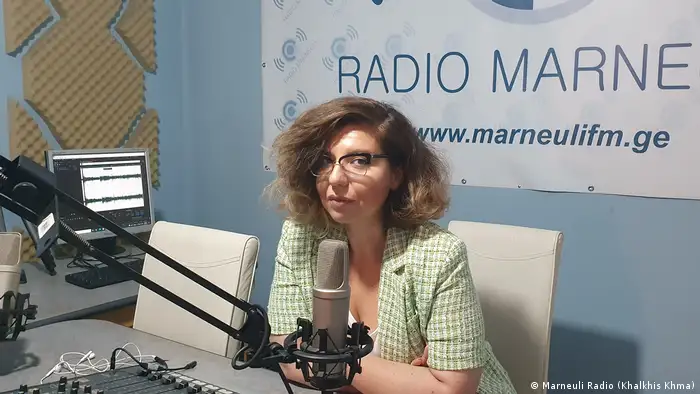 Kamila Mamedova is sitting in a radio studio in front of a mixing console and microphone. In the background, one wall is covered with soundproofing, and on the other hangs a banner with the name of the station Radio Marneuli and the website www.marneulifm.ge.