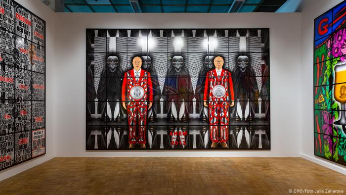 Gilbert & George artwork, two men dressed in red amid black-and-white masked figures. 