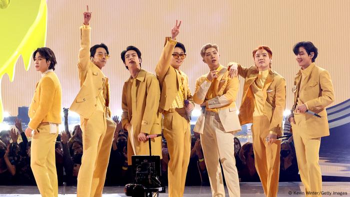 Members of the group BTS pose on stage, some saluting with a peace sign, all dressed in yellow. Members of the group BTS pose on stage, some saluting with a peace sign, all dressed in yellow.