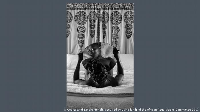 Photograph by Zanele Muholi showing a woman lying in bed and gazing at the camera lens through her own reflection in a mirror