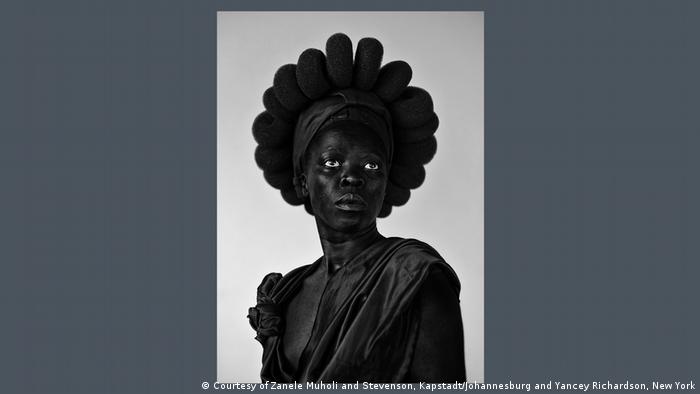 Photograph by Zanele Muholi showing a very dark female figure with bright eyes against a light background