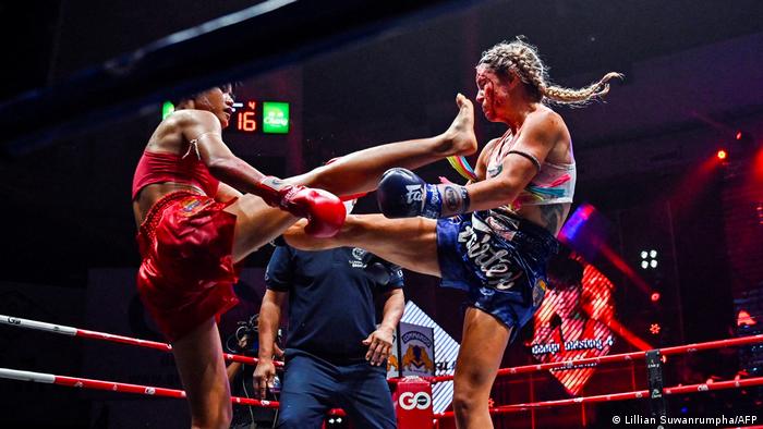 Two female boxers kick and bloody one another as a referee looks on. 