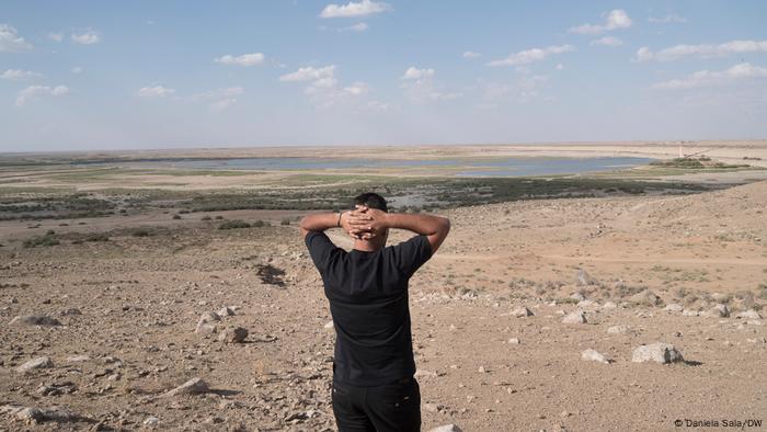 A man looks out over a severely shrunken lake, his arms folded behind his head