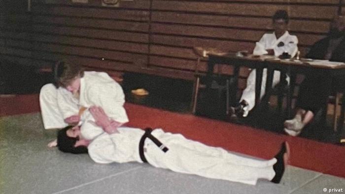 Blick in eine Sporthalle: Ein Mann im weißen Kampfanzug mit dunklem Gürtel liegt flach am Boden, eine Frau, ebenfalls im weißen Anzug hockt neben seinem Kopf, ihr angewinkeltes Bein liegt über seinen Schultern. Im Hintergrund sitzen zwei Männer an einem Tisch und schauen zu 