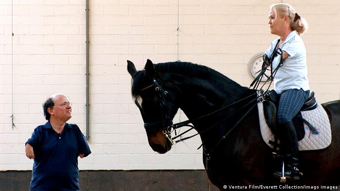 Eine Frau mit blondem Zopf sitzt auf einem schwarzen Pferd, das den Kopf mit weißer Blesse leicht geneigt hält. Ein Mann steht vor den beiden und schaut zu ihr auf. Beide haben kurze Arme. Aufnahme aus dem Film NoBody's Perfect von Niko von Glasow