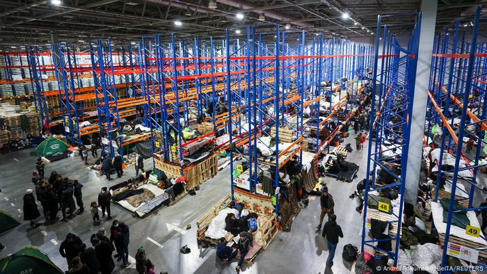 A wide shot showing a warehouse filled with cots and people
