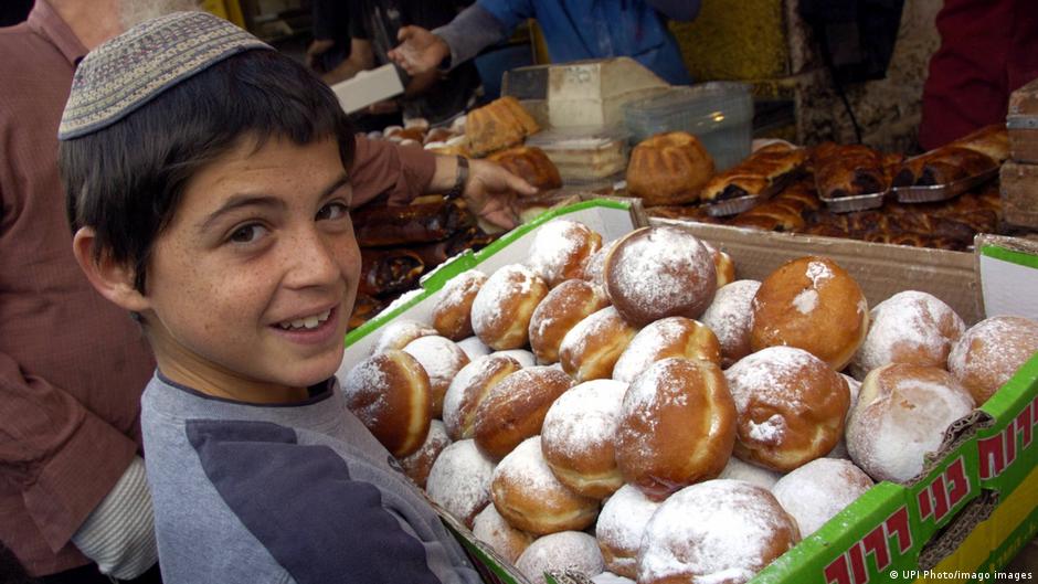 Ein Junge mit Kippa steht in Israel vor einem Marktstand und einer Kiste voller Krapfen.