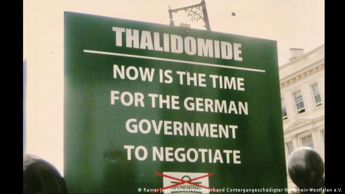 Blick auf ein grünes Plakat mit englischer Aufschrift, übersetzt lautet sie: Thalidomid: Jetzt muss die deutsche Regierung verhandeln. Es ist eine Aufnahme aus dem Film Stimmen gegen das Vergessen - Contergan