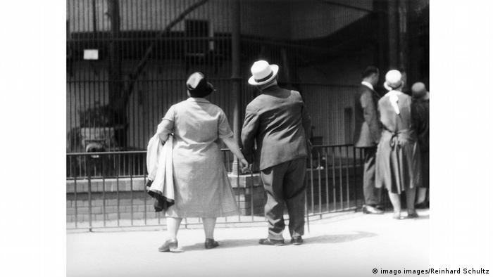 Photo by Tina Modotti: Overweight couple holding hands as they look at animals in a cage at the Berlin Zoological Garden.