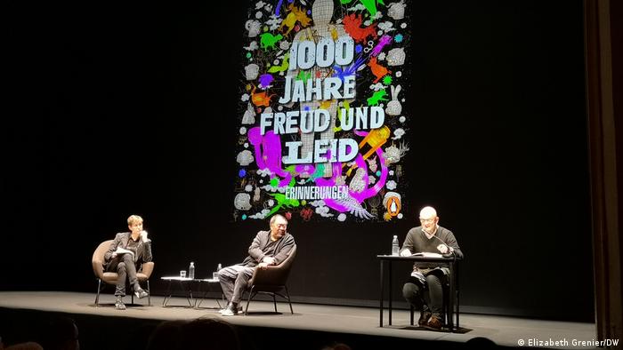 Buchpräsentation von AI Weiwei im Berliner Ensemble. Drei Männer sitzen auf einer Bühne, alle tragen schwarz, hinter ihnen wird das Buchcover auf eine Leinwand projiziert.