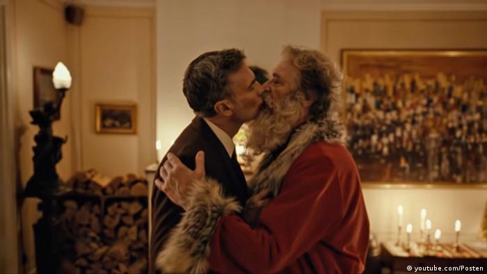 Ein Screenshot zeigt ein behagliches Wohnzimmer, links steht ein Mann in Anzugjacke, rechts der Weihnachtsmann. SIe küssen sich.