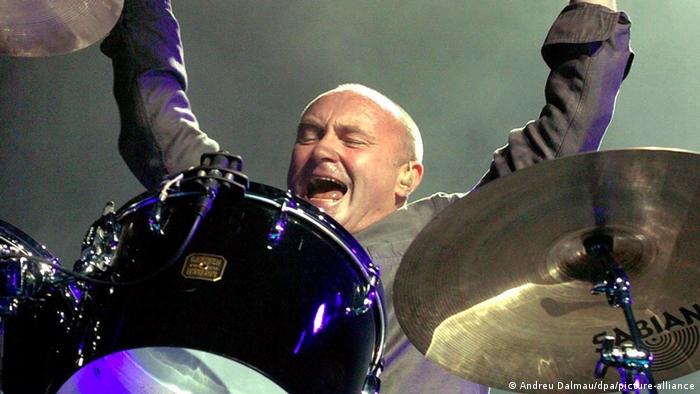 Phil Collins sitzt an den Drums und lacht und reißt die Arme nach oben.