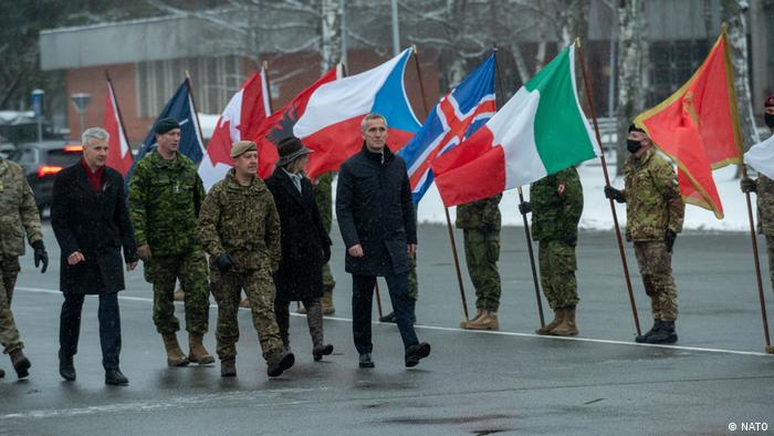 A group of men, some in unitorm, some in civilian clothing, walk past a rank of soldiers holding country flags of NATO members