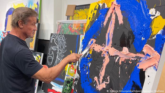 Schauspieler Sylvester Stallone steht in einem schwarzen T-Shirt vor einer Leinwand, auf der er malt.