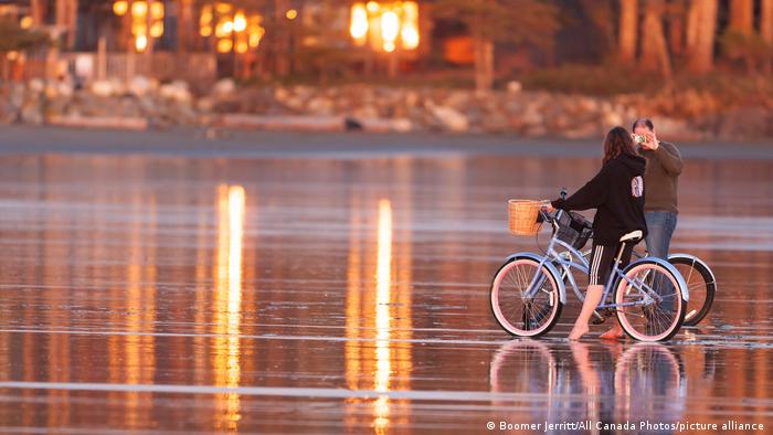 Ein Mann fotografiert eine Frau auf einem Fahrrad im Sonnenuntergang am Chesterman Beach in Tofino, Kanada 