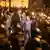 Pessoas em protesto noturno, rodeadas de policiais com capacete