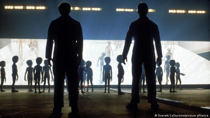 Still from a film showing two men with their backs to the camera looking at alien beings exiting a spaceship