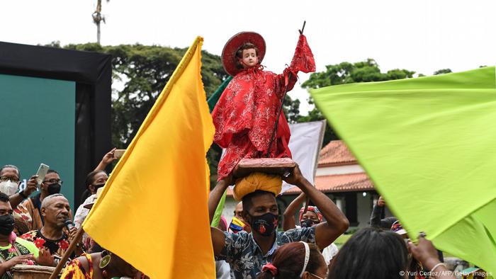 Un rassemblement de personnes avec des drapeaux et un homme tenant une figure d'un jeune saint Jean-Baptiste sur sa tête.