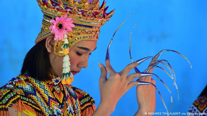A Nora performer in colorful dress and long metallic fingernails against a blue ground.