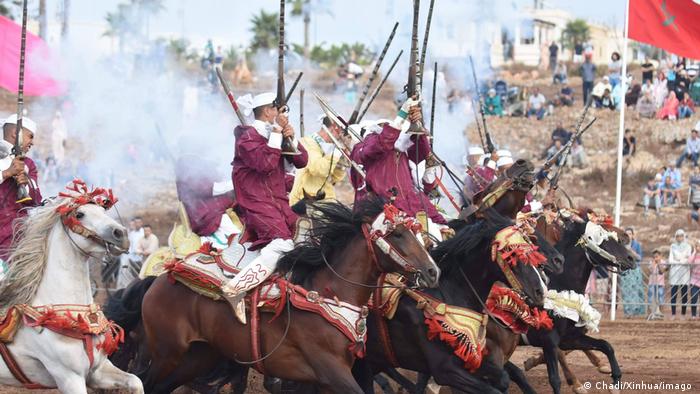 Horsemen in traditional Moroccan dress, carrying sabers and firing rifles as they ride.