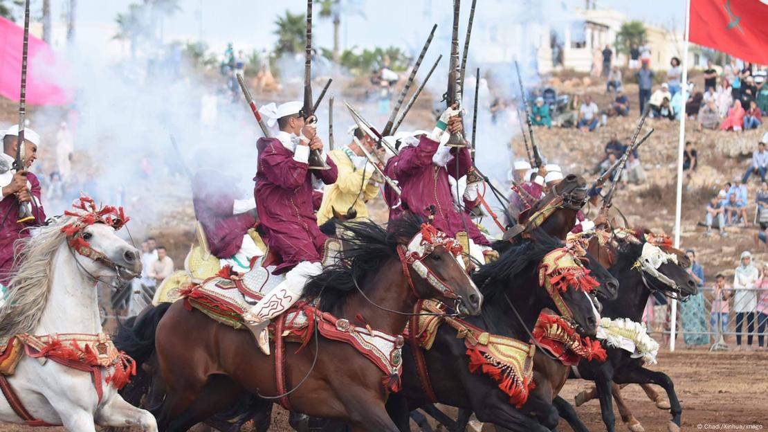 Horsemen in traditional Moroccan dress, carrying sabers and firing rifles as they ride.
