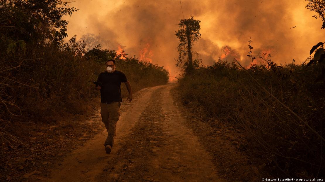 Homem foge de incêndio no Pantanal em 2020, usando máscara e correndo numa estrada de terra que vai até o horizonte; ao fundo da foto em tons marrons escuros, chamas e fumaça.