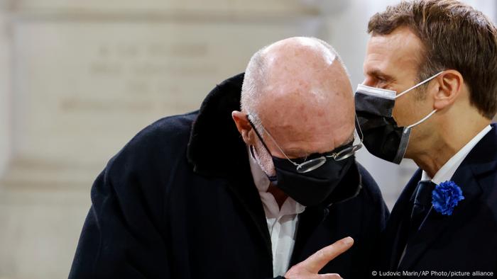 French President Emmanuel Macron (r) speaking with German artist Anselm Kiefer in 2020; both are wearing medical masks and Kiefer is leaning up to Macron's mouth to hear better.