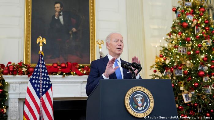 US President Joe Biden delivering remarks in the White House State Dining Room in front of an American flag, a Christmas tree and a portrait of Abraham Lincoln