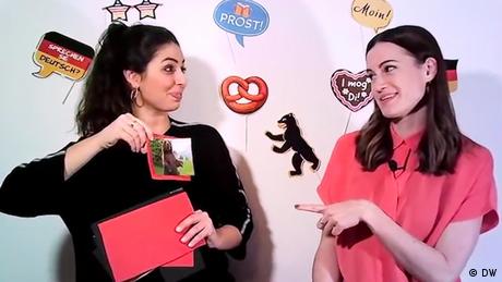 Someone showing a picture of a bear to another person pointing at her; there are different icons of German culture on the wall behind them, from a Bretzel to the Berlin bear.