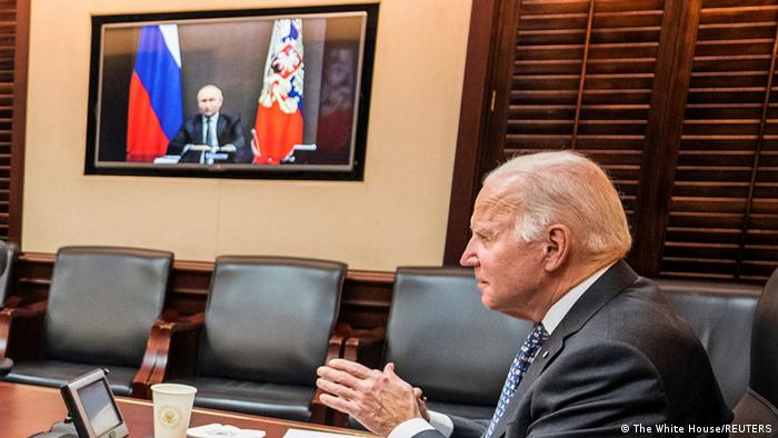 US president Joe Biden sitting at a table on a video call with Russian president Vladimir Putin, who is seen on a screen in the background