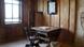 Foto mostra uma antiga mesa de uma antiga cadeira em uma sala de madeira. Há um quadro na parede e uma janela por onde entra luz. 