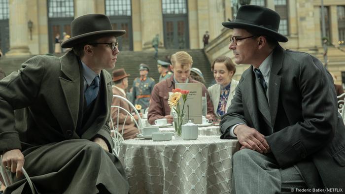 Film Still Munich - Edge of War Dois homens em casacos e chapéus sentados em uma pequena mesa um de frente para o outro 