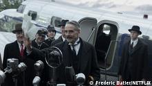 Film still Munich - the Edge of War, several men stand in front of microphones, behind them a small plane with an open door.
