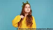 Little princess girl standing and pointing finger to herself, looking at camera with confidence, wearing yellow casual style sweater. Indoor studio shot isolated on blue background. || Modellfreigabe vorhanden