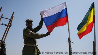 Un homme pose en tenue militaire en tenant le drapeau russe à côté d'un drapeau éthiopien