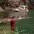 Funcionário do Corpo de Bombeiros, vestido com uniforme vermelho, olha sobre o lago de Furnas enquanto embarcação, ao fundo, realiza busca por vítimas da queda de um bloco de rocha de um paredão no local. Do lado direito do lago verde, é possível ver o pedaço de uma cachoeira.