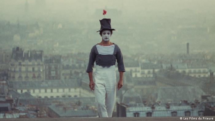 Marcel Marceau steht als Clown Bip verkleidet mit Ringelshirt und weißem Gesicht auf einem Dach. Hinter ihm sind die Häuser von Paris zu sehen.