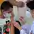 De máscara, menino olha para o braço esquerdo descoberto enquanto funcionária vestida com jaleco branco e máscara aplica vacina no garoto em São Paulo.