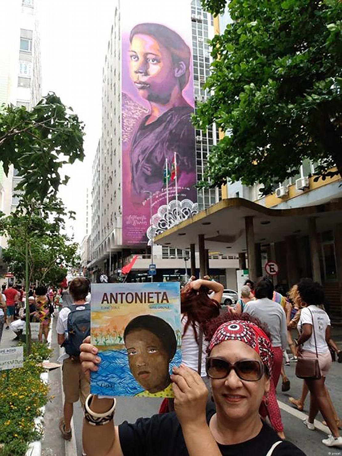 A professora Eliane Debus em frente ao mural em homenagem a Antonieta, em Florianópolis, segurando seu livro infantil sobre a educadora