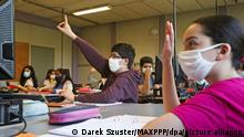 Schüler mit Masken sitzen in einem Klassenraum, einige zeigen auf 