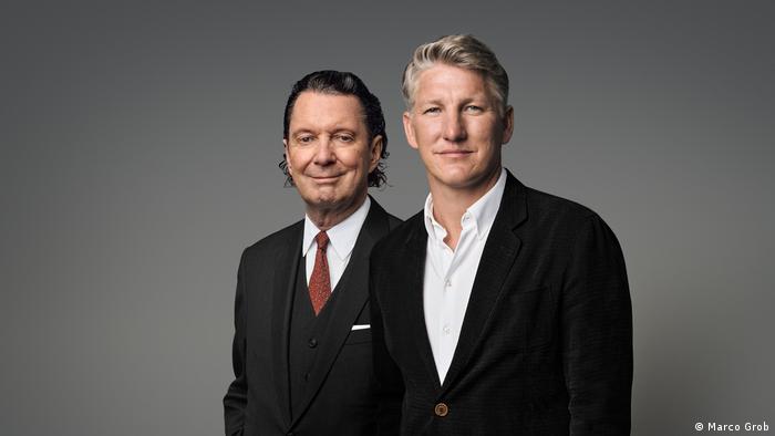 Martin Suter and Bastian Schweinsteiger look into camera, standing side by side, wearing dark suits 