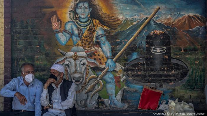 Mural painting of Shiva on a wall, two men sitting in front of it wearing masks
