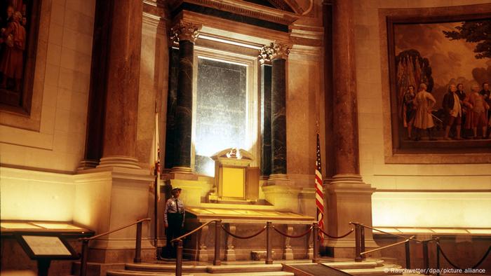 Grand hall avec des colonnes à gauche et à droite d'un document inscrit dans du verre, bouclé, flanqué d'un garde et du drapeau américain.
