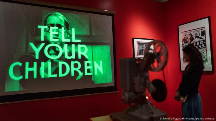 Woman standing beside a projector looks at a screen on a wall showing a person and the words in caps, Tell your Children, at the 'Museum of Weed' in Los Angeles